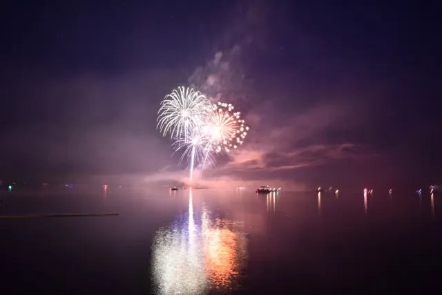 White and pink fireworks reflected across Lake Arbutus, with boats on the water.