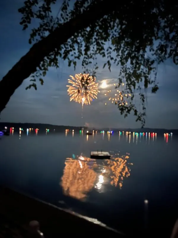 Golden fireworks and their lake reflection framed by shoreline trees.